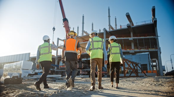 A group of four construction workers walk towards a construction site. They are wearing yellow and orange vests. It is early morning.