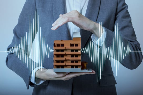 Man in a suit holds a small model building while white vibration waves stream from the building's sides.